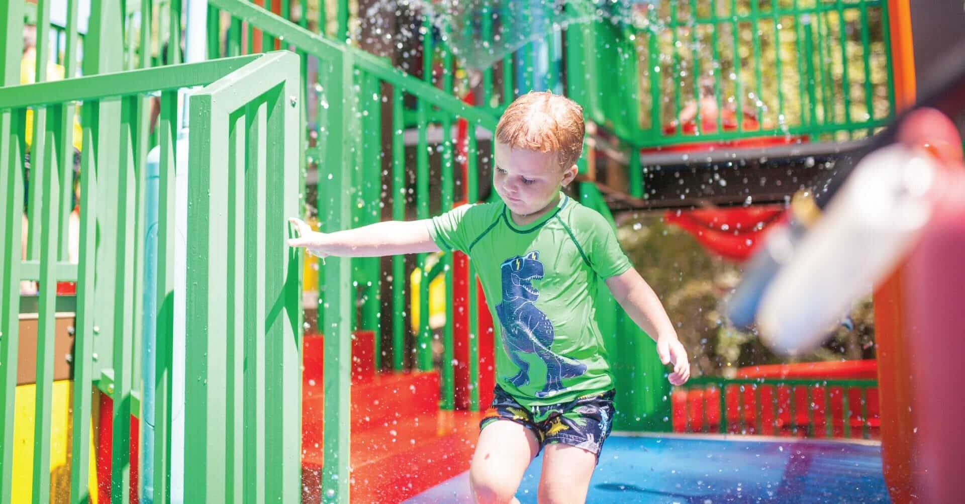 A little boy exits the water play structure by Interactive Play, water sprays in the background.