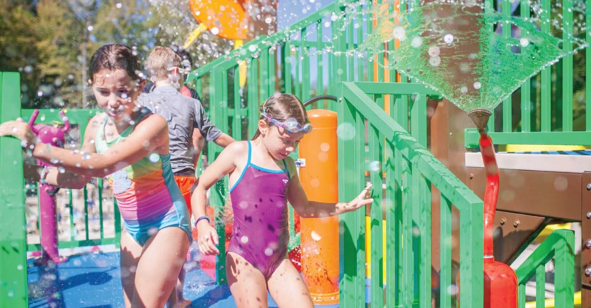 A little girl exits the aquatic play unit and is sprayed with a cascading water spray by the fanatic water feature next to her.