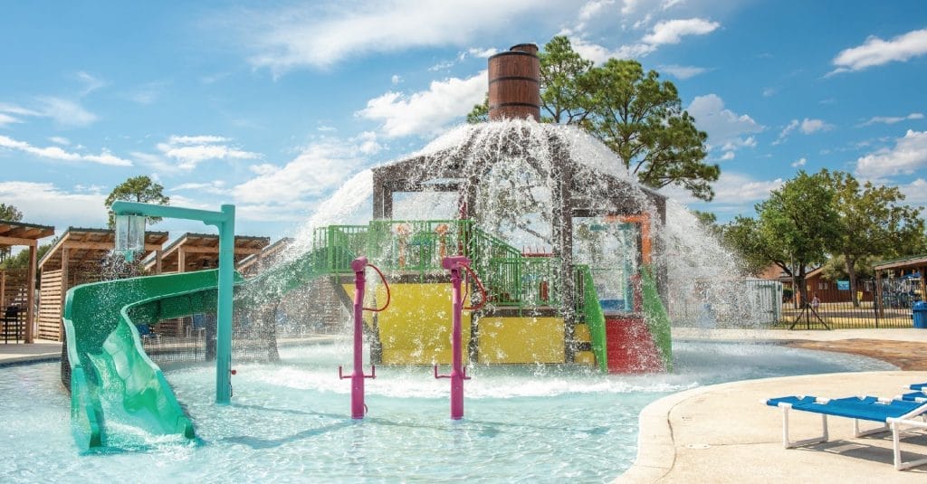 A photo of the aquatic play structure by Interactive play with the power plunge feature releasing a large volume of water cascading over the structure and onto the pool deck at the RV resort in Waller, Texas.